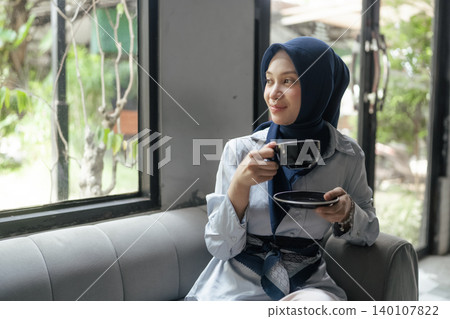 Smiling woman in hijab holding a cup and saucer while sitting on a sofa near a bright window Smiling woman in hijab holding a cup and saucer while sitting on a sofa near a bright window 140107822