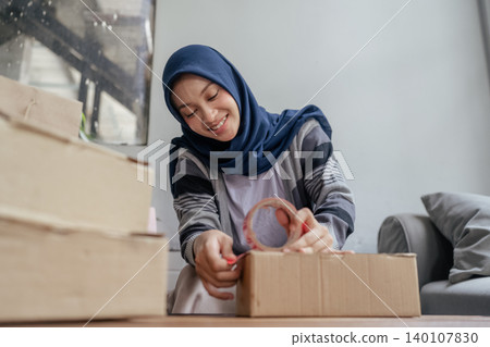 Smiling woman in hijab sealing a cardboard box while packing belongings at home Smiling woman in hijab sealing a cardboard box while packing belongings at home 140107830