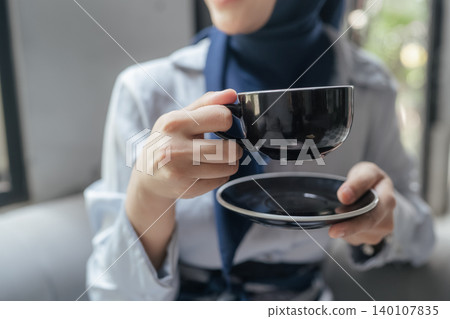 Person holding a black teacup and saucer in a softly lit indoor setting Person holding a black teacup and saucer in a softly lit indoor setting 140107835