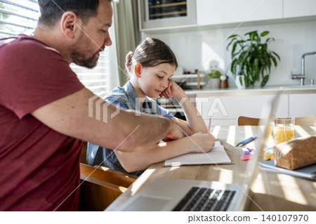 Daughter studying at table as dad works remotely on laptop nearby. 140107970