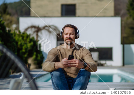Man sitting by swimming pool listening to music with headphones. Man sitting by swimming pool listening to music with headphones. 140107996
