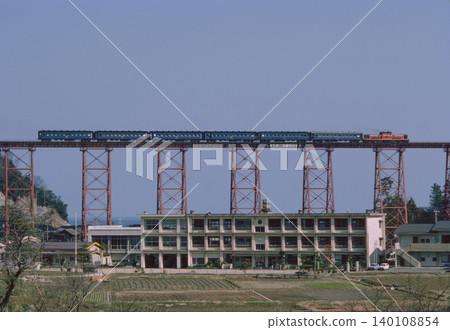 A DD51 locomotive and six old-style passenger cars cross the Amarube Viaduct overlooking the Sea of Japan. A railway heritage trestle bridge. 1986 archive. A DD51 locomotive and six old-style passenger cars cross the Amarube Viaduct overlooking the Sea of Japan. A railway heritage trestle bridge. 1986 archive. 140108854