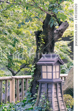 A vertical composition of a stone lantern in front of a large tree. A vertical composition of a stone lantern in front of a large tree. 140108920