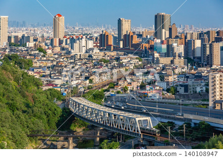 A JR Takarazuka Line train runs across a railway bridge over the Mukogawa River, with the cityscape of Takarazuka, Hyogo Prefecture, and the Osaka Plain in the background. 140108947