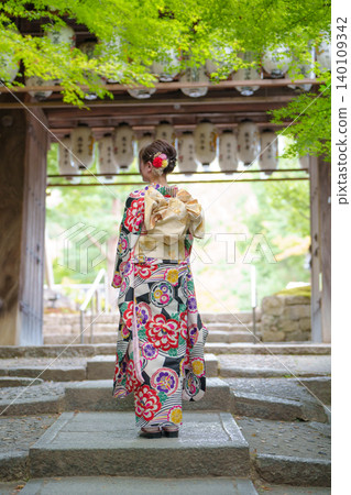 The back view of a woman in a kimono standing on stone steps beneath a traditional wooden gate. Kyoto, Japan. The back view of a woman in a kimono standing on stone steps beneath a traditional wooden gate. Kyoto, Japan. 140109342