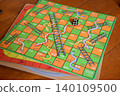 A close-up overhead angled view of a classic Snakes and Ladders board game laid out on a warm brown wooden table surface. 140109500