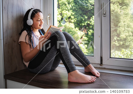 Young girl reading book and listening music by window Young girl reading book and listening music by window 140109783