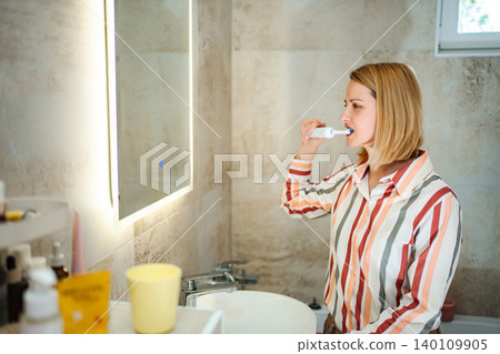 Woman brushing teeth with electric toothbrush reflecting in mirror Woman brushing teeth with electric toothbrush reflecting in mirror 140109905