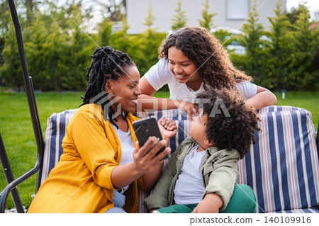 Black mother and children connecting with phone in backyard 140109916