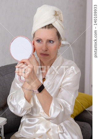 Woman in a silk robe looks in a handheld mirror while preparing for the day in a cozy indoor setting Woman in a silk robe looks in a handheld mirror while preparing for the day in a cozy indoor setting 140109933