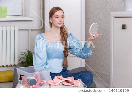 Young woman examining herself in a handheld mirror while sitting in a cozy indoor space 140110109