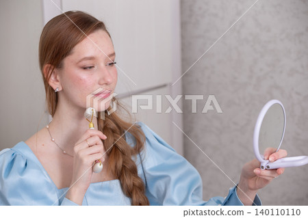 Young woman in blue dress using facial roller while looking in mirror in bright indoor setting during daytime 140110110