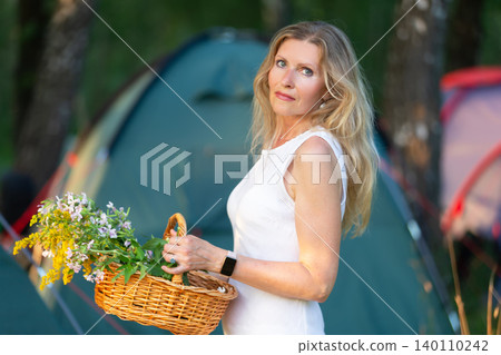 Woman holding a basket of flowers in a campsite surrounded by tents during sunset in a forest clearing 140110242