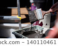 Hand holds paintbrush with dark paint dripping over tray, with bucket and ladder in background 140110461
