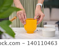 Woman prepares yellow plant pot on gardening table with bowls, scattered soil and plant leaves nearby 140110463