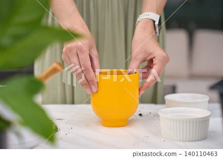 Woman prepares yellow plant pot on gardening table with bowls, scattered soil and plant leaves nearby 140110463