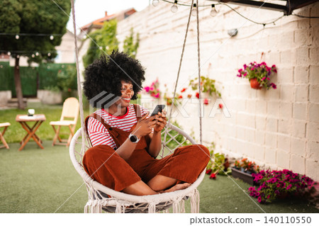 Woman relaxing in hanging chair browsing smartphone in backyard garden Woman relaxing in hanging chair browsing smartphone in backyard garden 140110550