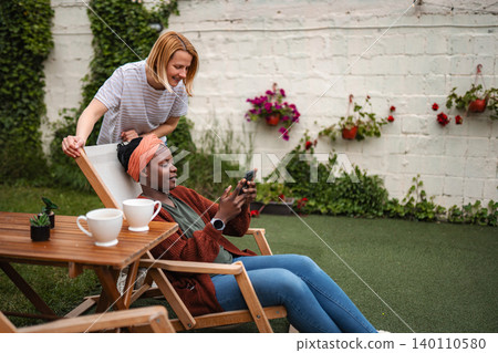 Friends relaxing together in backyard enjoying coffee and smartphone Friends relaxing together in backyard enjoying coffee and smartphone 140110580