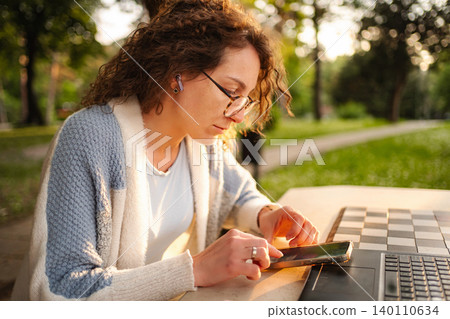 Young woman working remotely with smartphone and laptop in park at sunset Young woman working remotely with smartphone and laptop in park at sunset 140110634