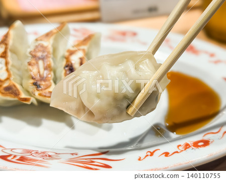 A fragrant pan-fried dumpling eaten at a ramen shop. A close-up of a dumpling held with chopsticks. Dipping it in the sauce and eating it. A fragrant pan-fried dumpling eaten at a ramen shop. A close-up of a dumpling held with chopsticks. Dipping it in the sauce and eating it. 140110755