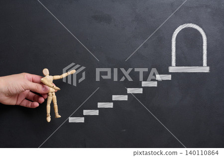 A woman's hand places a wooden mannequin on a chalk-drawn staircase leading to an open door on a blackboard. A woman's hand places a wooden mannequin on a chalk-drawn staircase leading to an open door on a blackboard. 140110864