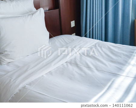 White bed linens with neatly arranged pillows on a hotel bed, blue curtains partially visible in the background, creating a serene indoor atmosphere White bed linens with neatly arranged pillows on a hotel bed, blue curtains partially visible in the background, creating a serene indoor atmosphere 140111062