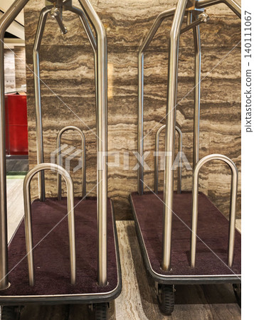 Hotel luggage carts with metal frames and plush fabric bases positioned against a textured stone wall in a modern lobby area Hotel luggage carts with metal frames and plush fabric bases positioned against a textured stone wall in a modern lobby area 140111067