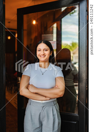 Smiling woman standing with arms crossed at entrance of coffee shop 140111242
