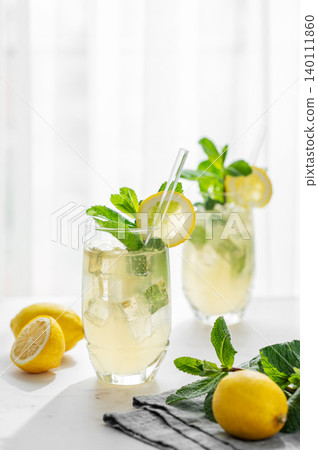 A summer drink with lemon, mint, and ice in a two glasses against a sunny window background. A summer drink with lemon, mint, and ice in a two glasses against a sunny window background. 140111860