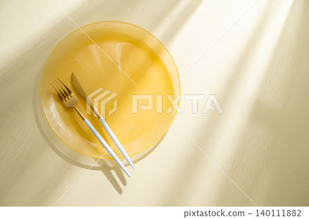An empty yellow plate with a fork, knife on a light background with sunlight. An empty yellow plate with a fork, knife on a light background with sunlight. 140111882
