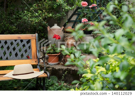 Wooden garden bench with straw hat resting on it, surrounded by vibrant flowers and potted plants in a lush garden setting Wooden garden bench with straw hat resting on it, surrounded by vibrant flowers and potted plants in a lush garden setting 140112029