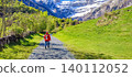 Hiker in red jacket walks along a gravel path in the French Pyrenees with snow-capped mountains and lush greenery in the background 140112052