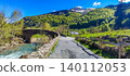 Scenic view of a winding road leading to a stone bridge over a river in the Pyrenees mountains with lush greenery and blue sky 140112053