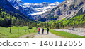 Group of hikers walking along a dirt path in the French Pyrenees, surrounded by lush greenery and snow-capped mountains in the background 140112055