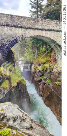 Stone arch bridge spanning a rocky gorge in the French Pyrenees, with rushing water and lush greenery surrounding the scene 140112062