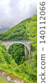 Stone arch bridge spanning a lush green valley in the French Pyrenees, surrounded by dense trees and misty mountains in the background 140112066