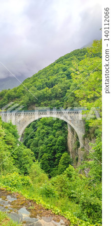 Stone arch bridge spanning a lush green valley in the French Pyrenees, surrounded by dense trees and misty mountains in the background Stone arch bridge spanning a lush green valley in the French Pyrenees, surrounded by dense trees and misty mountains in the background 140112066