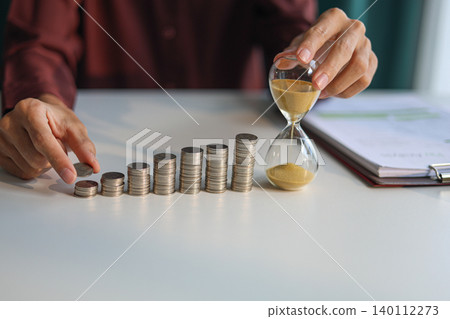 Time and money management concept. Rising stack of coins and running sand clock on the table. Growing of income, saving or return investment through time. Time and money management concept. Rising stack of coins and running sand clock on the table. Growing of income, saving or return investment through time. 140112273