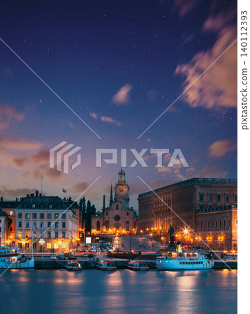 night view overlooking the illuminated historic architecture of Gamla Stan reflecting in the calm waters of Stockholm, Sweden, under a starry night sky 140112393