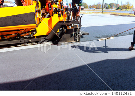 Workers use asphalt paving machines to lay asphalt on parking lot while another worker smooths surface with tool. 140112481