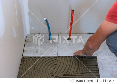 Worker lays tiles in bathroom while adjusting plumbing pipes space is under renovation home Worker lays tiles in bathroom while adjusting plumbing pipes space is under renovation home 140112507