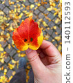 Close-up photo of a hand holding a small colorful autumn leaf with smile pattern. Cobblestone covered yellow leaves on background. Cozy autumn atmosphere, fall mood, happiness, or mindfulness 140112593