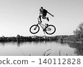 Black and white photo of a person performing a high jump with a bicycle over a river. The boy is captured mid-air in an acrobatic pose against a clear sky and calm water. Freedom, adventure, youth 140112828