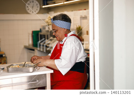 Senior Black woman happily preparing fresh handmade pastries in a bright commercial kitchen setting 140113201