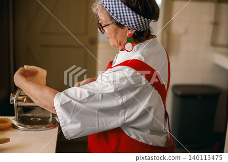 Senior Caucasian woman handcrafting fresh pasta with a machine in a bright domestic kitchen Senior Caucasian woman handcrafting fresh pasta with a machine in a bright domestic kitchen 140113475
