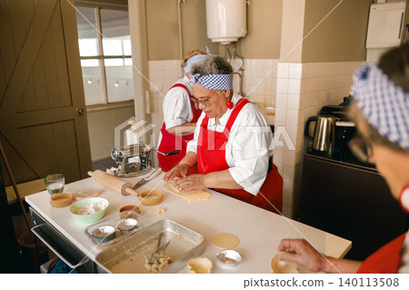Senior multiracial women rolling dough for baking in a bright kitchen Senior multiracial women rolling dough for baking in a bright kitchen 140113508