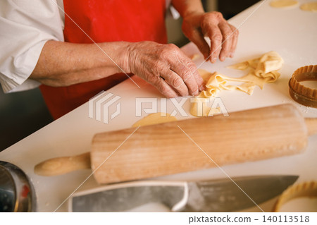 Senior Caucasian woman hands preparing homemade dough with rolling pin in bright kitchen bakery Senior Caucasian woman hands preparing homemade dough with rolling pin in bright kitchen bakery 140113518