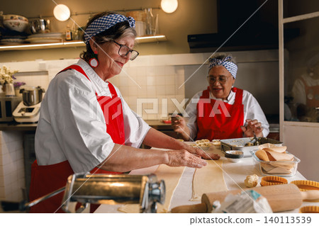 Active Caucasian and Black senior women rolling dough in a bright commercial kitchen setting Active Caucasian and Black senior women rolling dough in a bright commercial kitchen setting 140113539