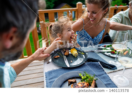Family eating at barbecue party dinner on patio, little girl eating roasted corn and enjoying it. Family eating at barbecue party dinner on patio, little girl eating roasted corn and enjoying it. 140113578