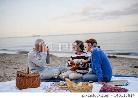 Grandparents enjoying beach picnic with teenage granddaughter playing ukulele by sea. Grandparents enjoying beach picnic with teenage granddaughter playing ukulele by sea. 140113614